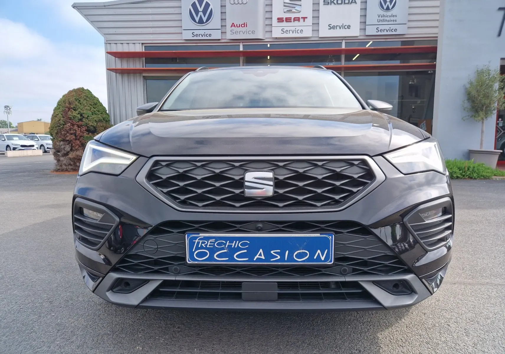 Vue de face d’un SEAT Ateca noir 2023 avec calandre hexagonale et plaques d’immatriculation « Fréchic Occasion » devant un garage multimarques.