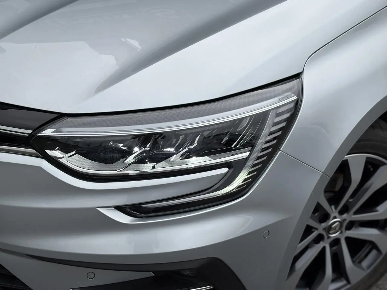 Gros plan sur le phare avant droit et la jante alliage d'une Renault Mégane Break 2024 gris Highland.
