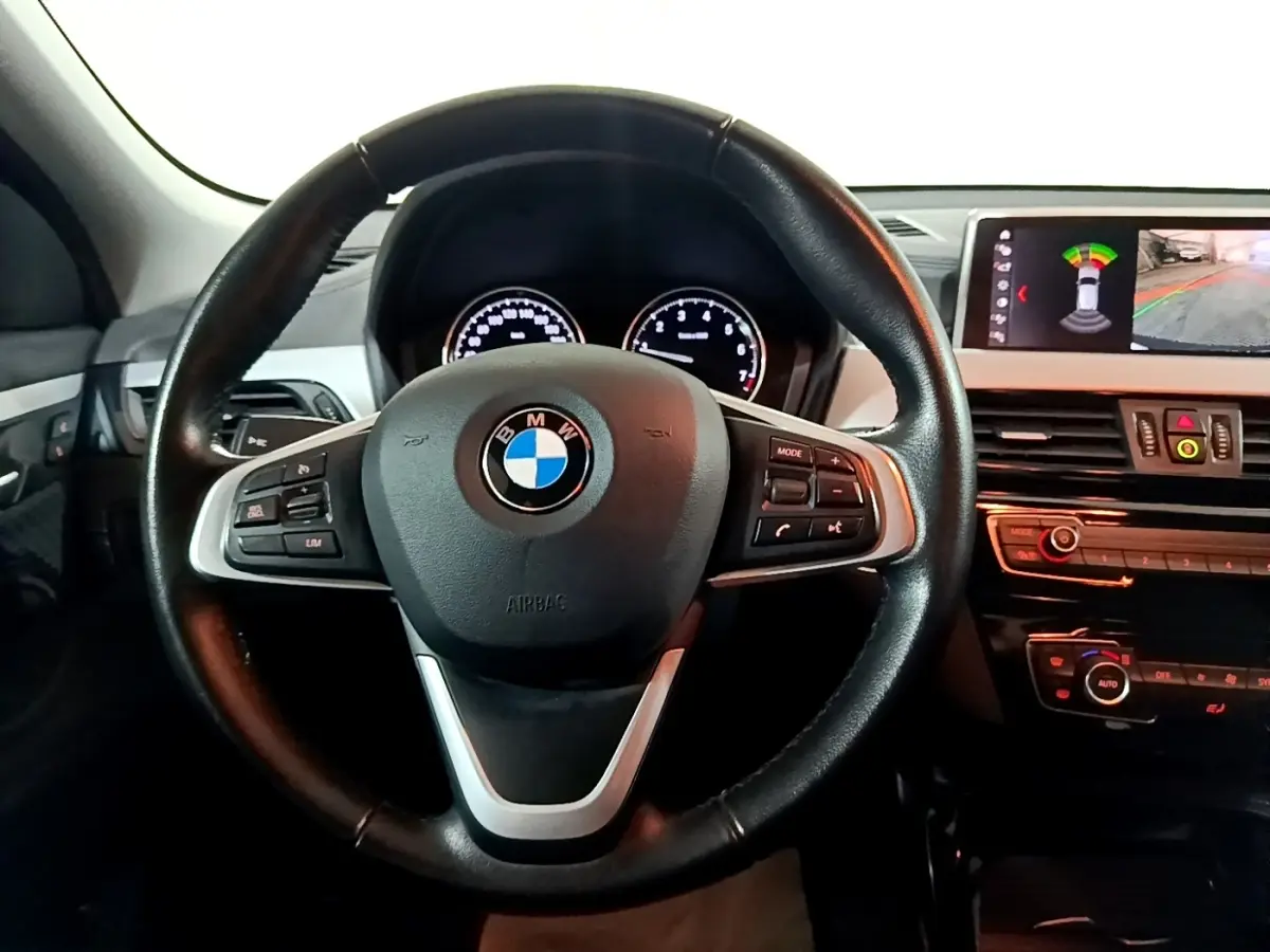 Vue intérieure centrée sur le volant cuir noir de la BMW X2 avec tableau de bord et écran d'aide au stationnement.