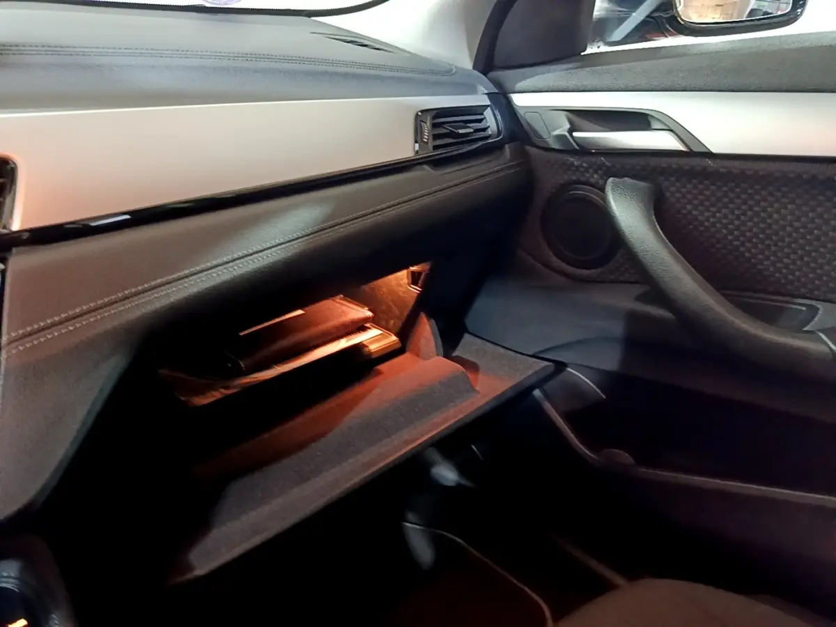 Intérieur BMW X2 sDrive18i 2023 côté passager, boîte à gants ouverte avec éclairage ambiant orange.