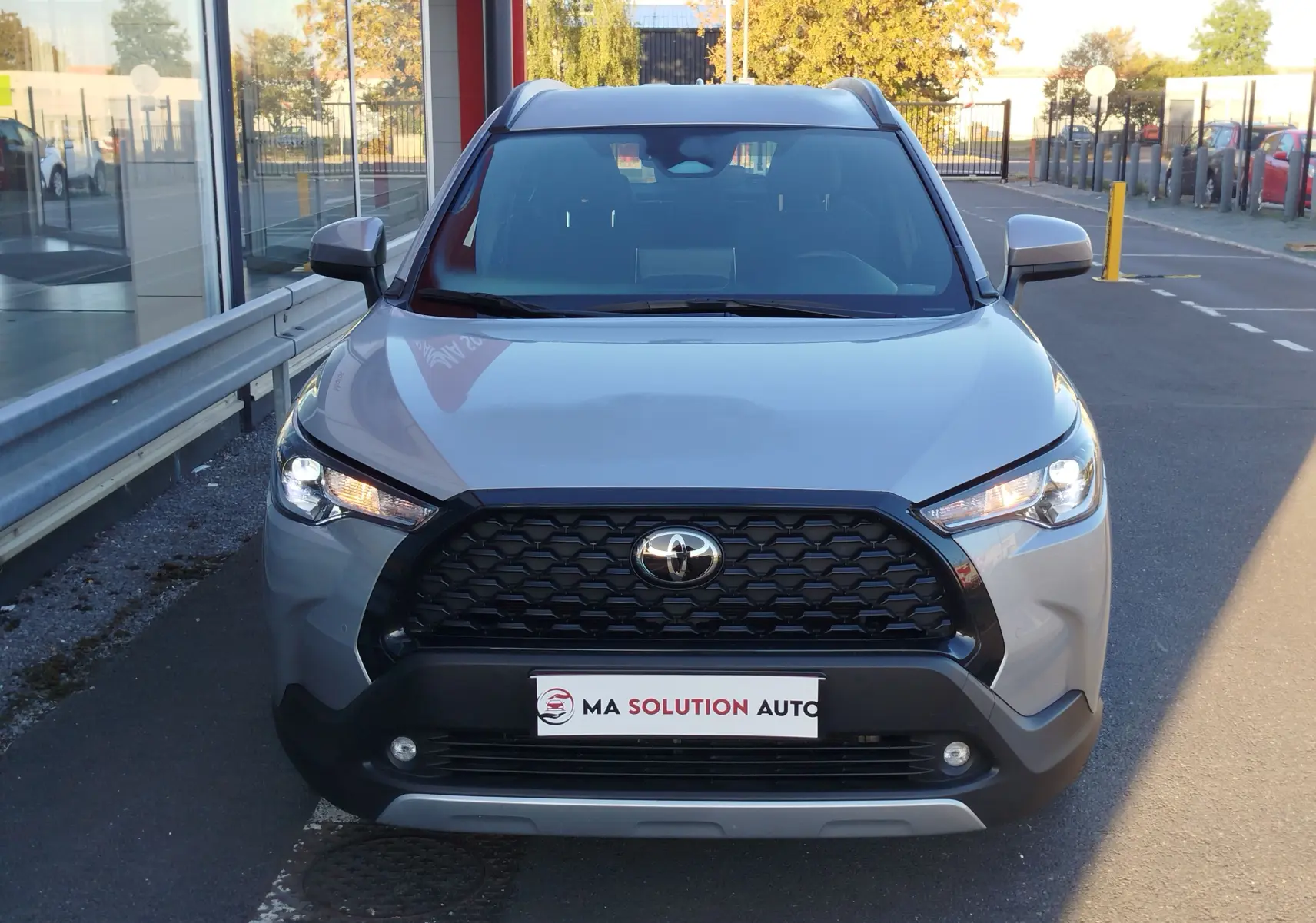 Vue frontale d'un Toyota Corolla Cross gris minéral avec calandre noire et phares LED allumés, stationné devant un bâtiment.