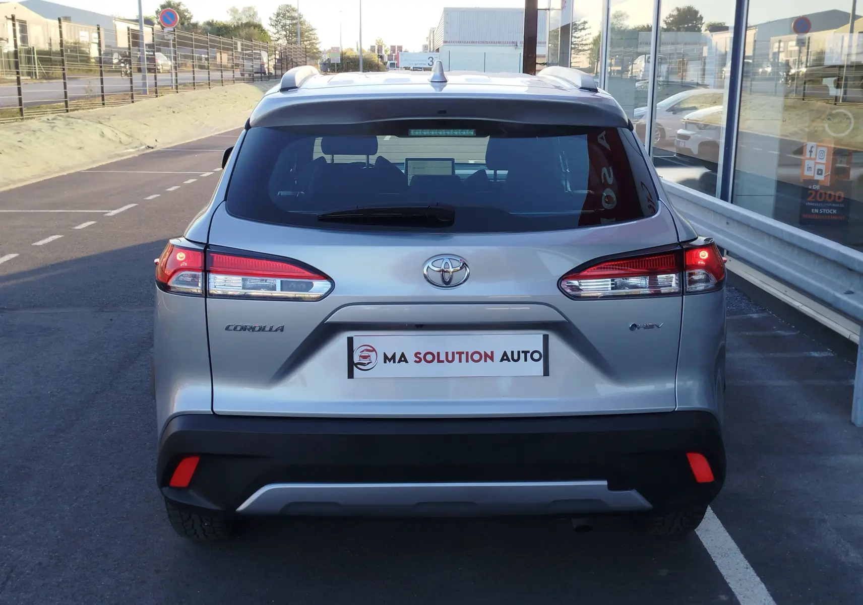 Vue arrière d'un Toyota Corolla Cross gris minéral 2025 avec feux LED et logo hybride visible sur un parking.