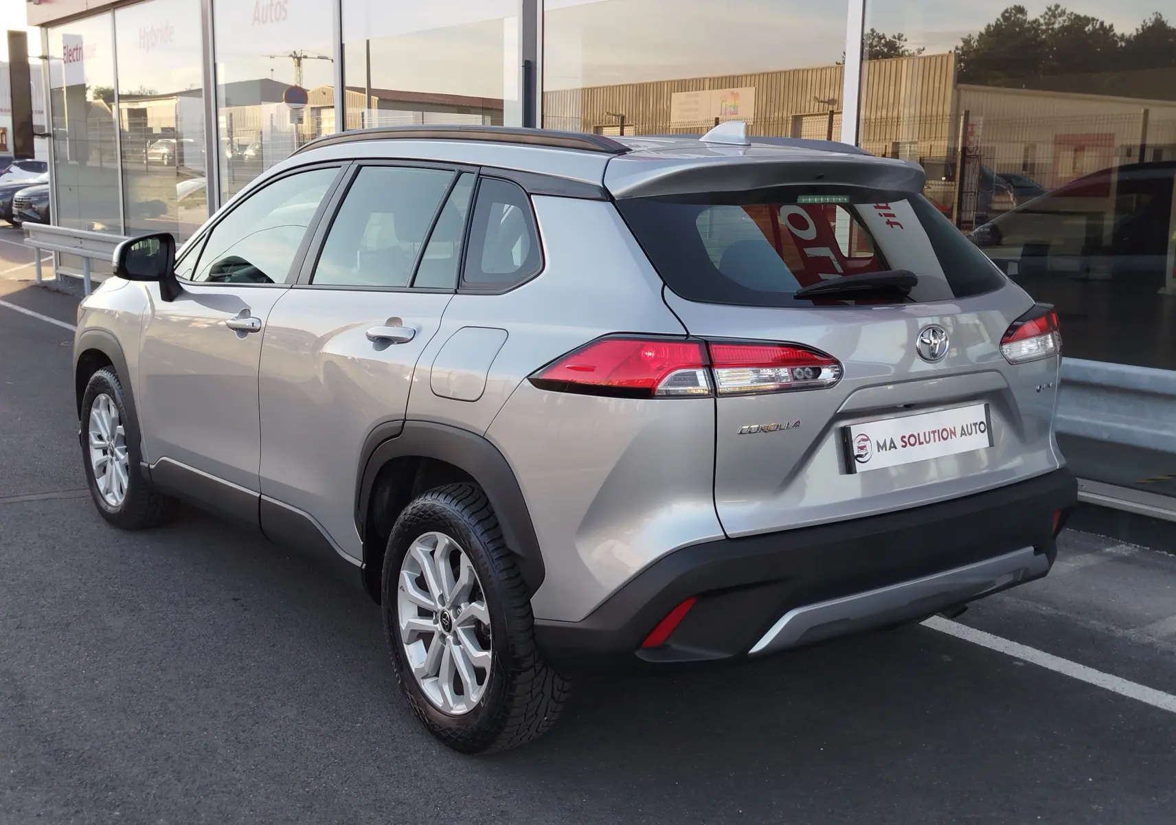 Vue 3/4 arrière droite d'un Toyota Corolla Cross gris minéral avec jantes alliage et feux arrière LED, stationné devant un showroom.