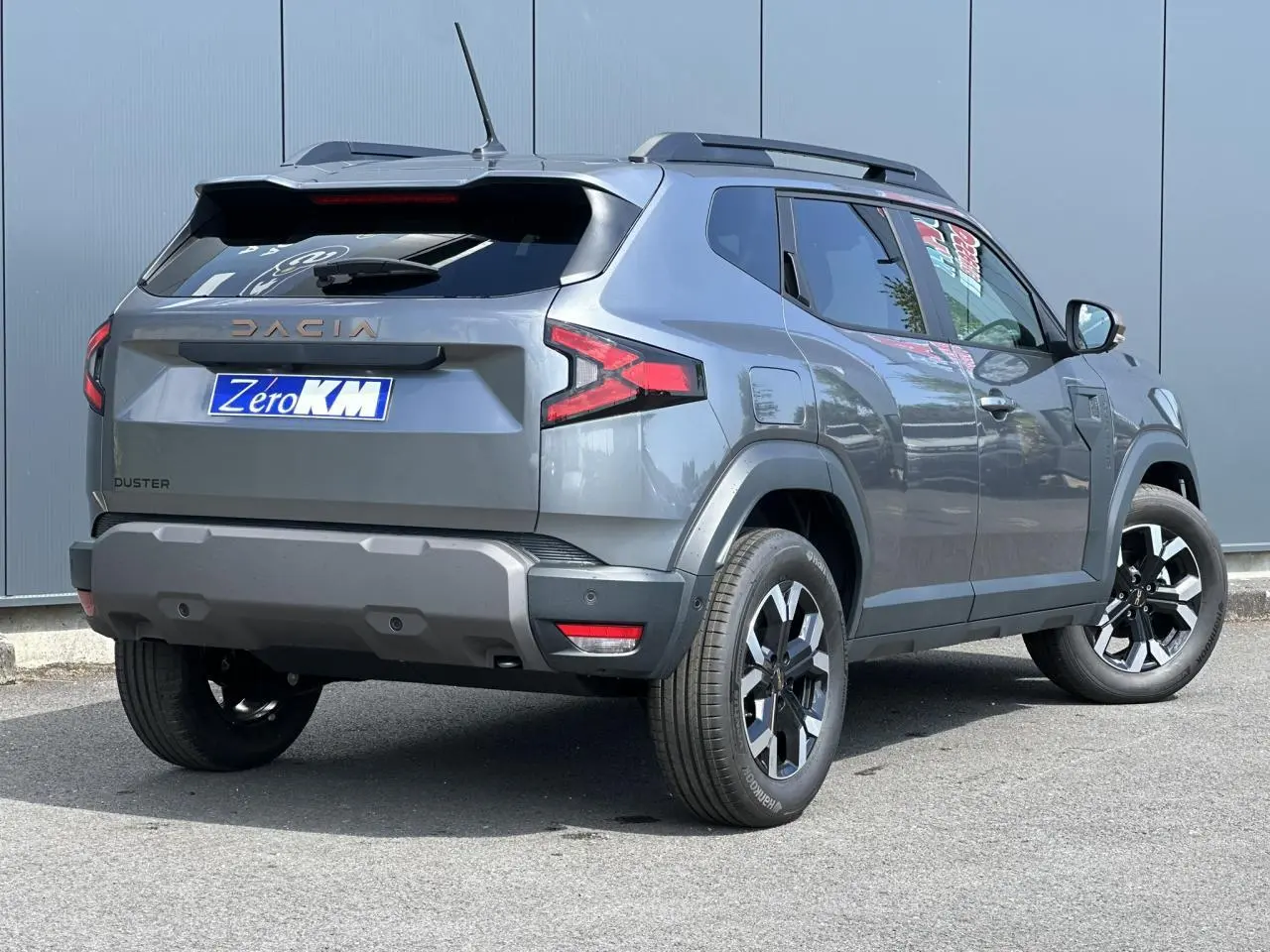 Vue 3/4 arrière droite du Dacia Duster 2025 gris schiste avec jantes alliage et barres de toit noires.