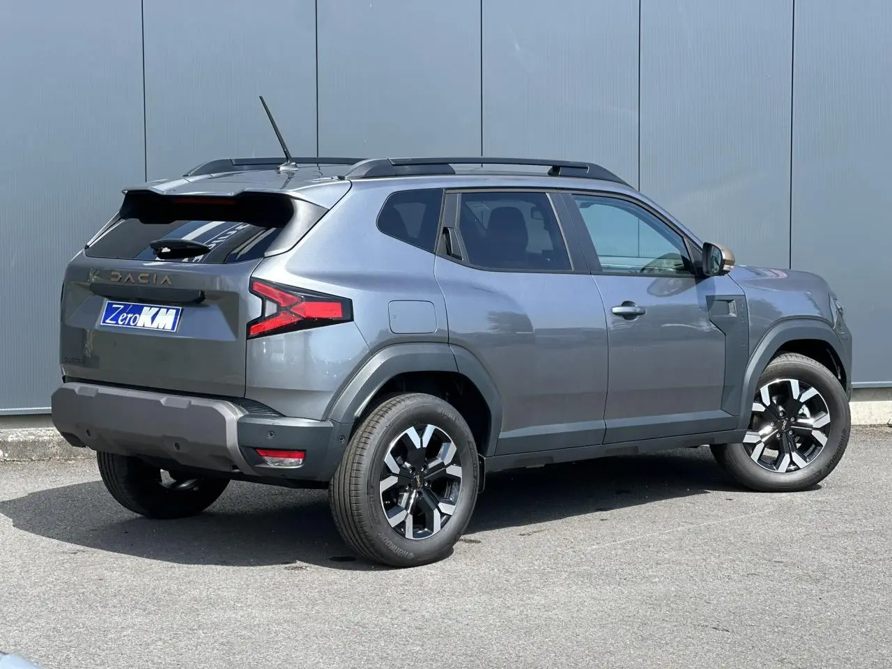 Dacia Duster Gris Schiste en 3/4 arrière droit, avec barres de toit noires et jantes alliage 17 pouces.