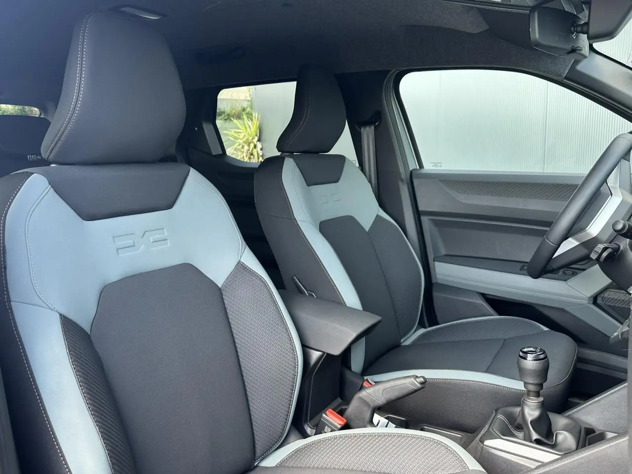 Vue intérieure côté conducteur du Dacia Duster 2025, sièges bi-ton gris clair et foncé avec logo embossé sur appuie-tête.