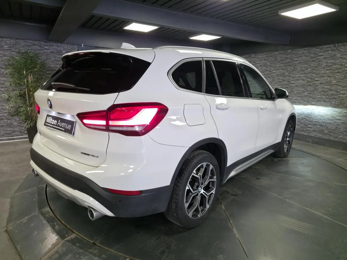 BMW X1 sDrive 18d blanc vue 3/4 arrière droit, avec jantes alliage 18 pouces et sorties d’échappement chromées.