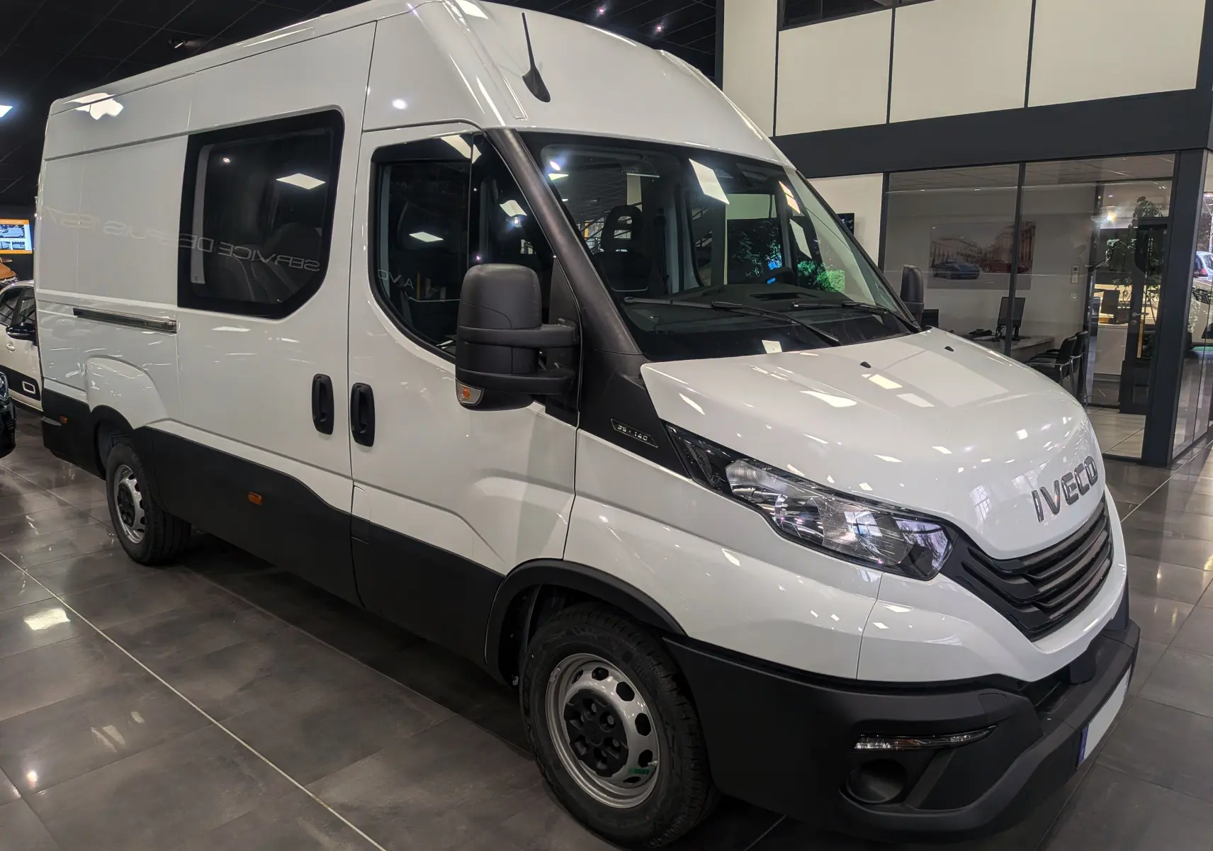 Fourgon Iveco Daily blanc vu en 3/4 avant droit dans un showroom avec vitres teintées et pare-chocs noirs.