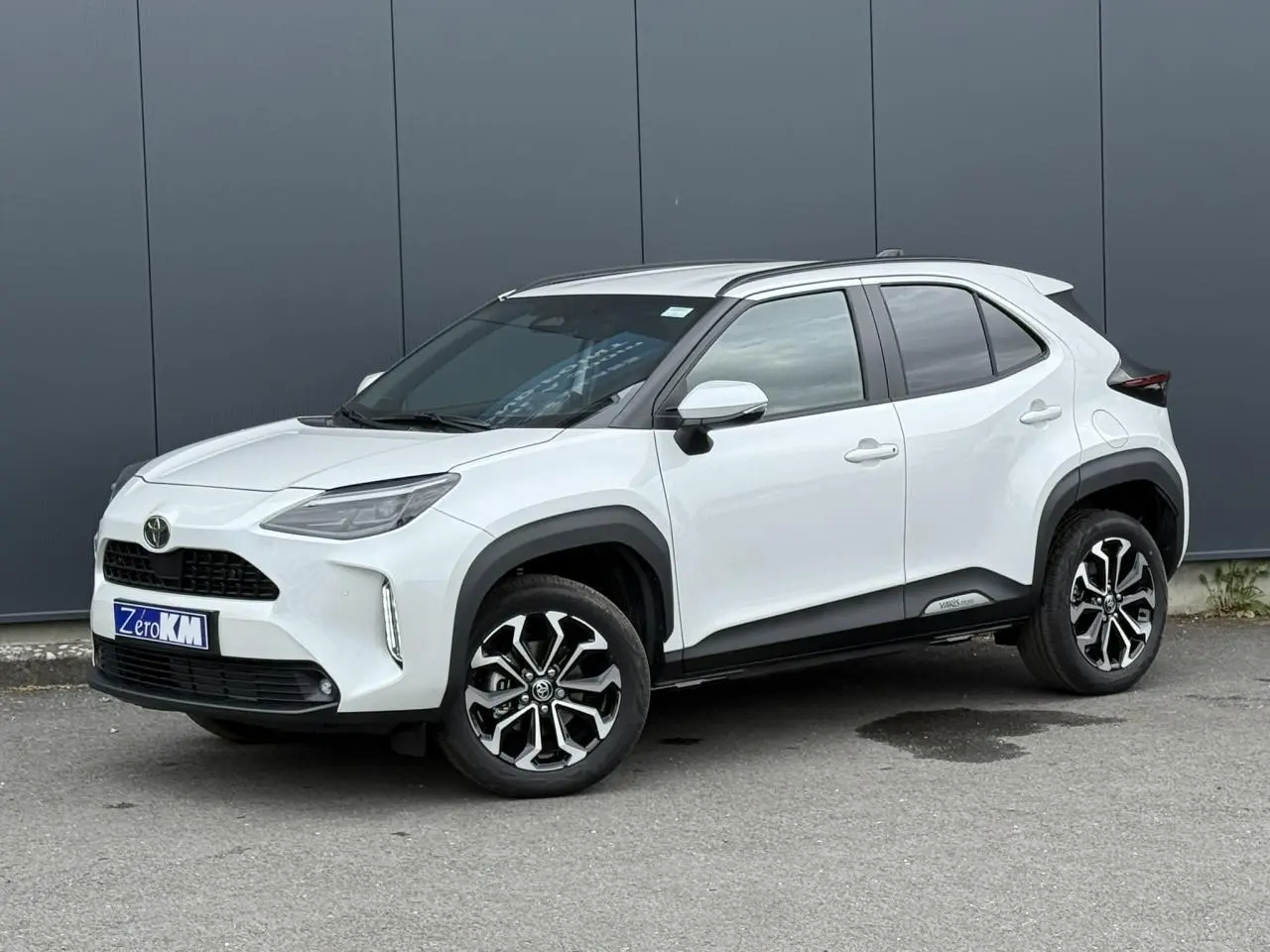 Toyota Yaris Cross blanc lunaire vue 3/4 avant droit, avec jantes alliage et protections noires contrastées.
