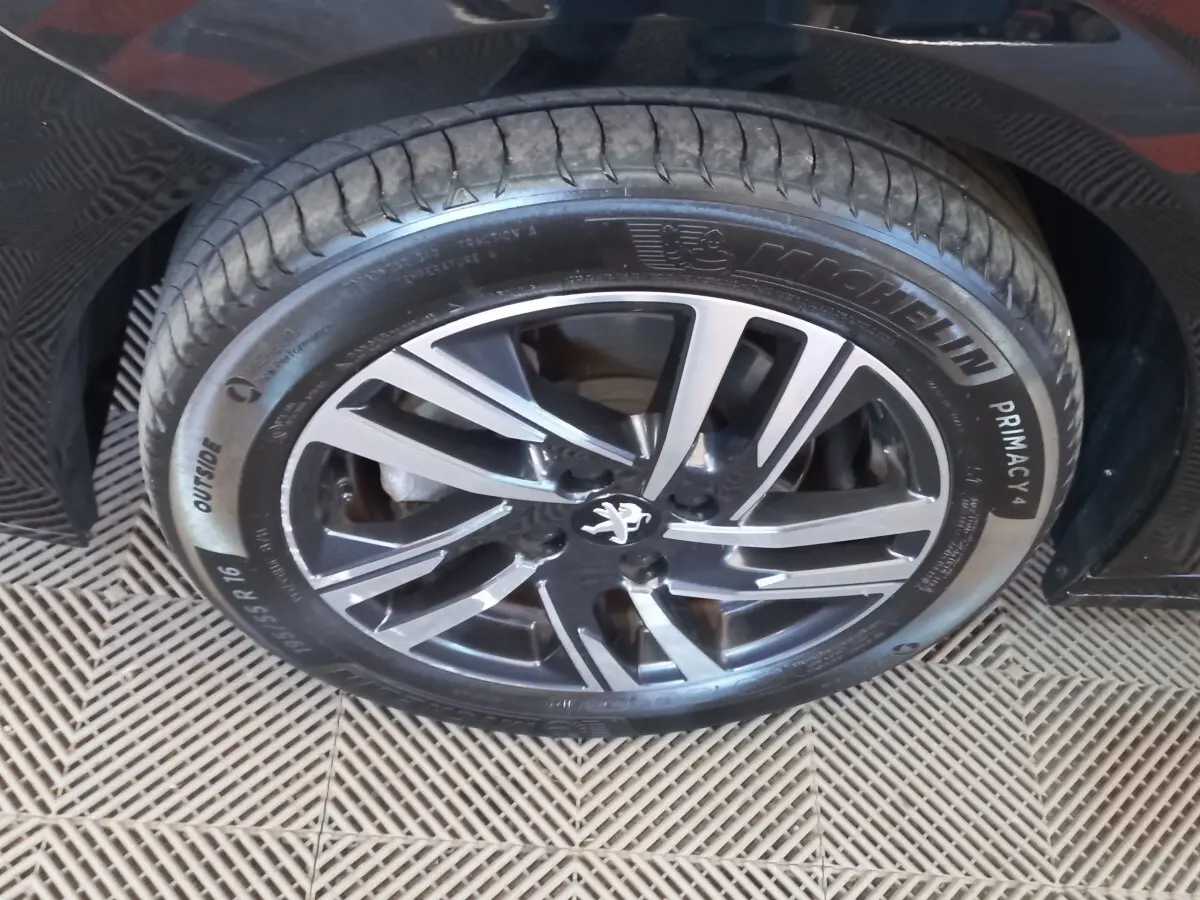 Gros plan sur la jante alliage noire et argentée avec logo Peugeot, montée sur un pneu Michelin Primacy 4, voiture noire visible en arrière-plan.
