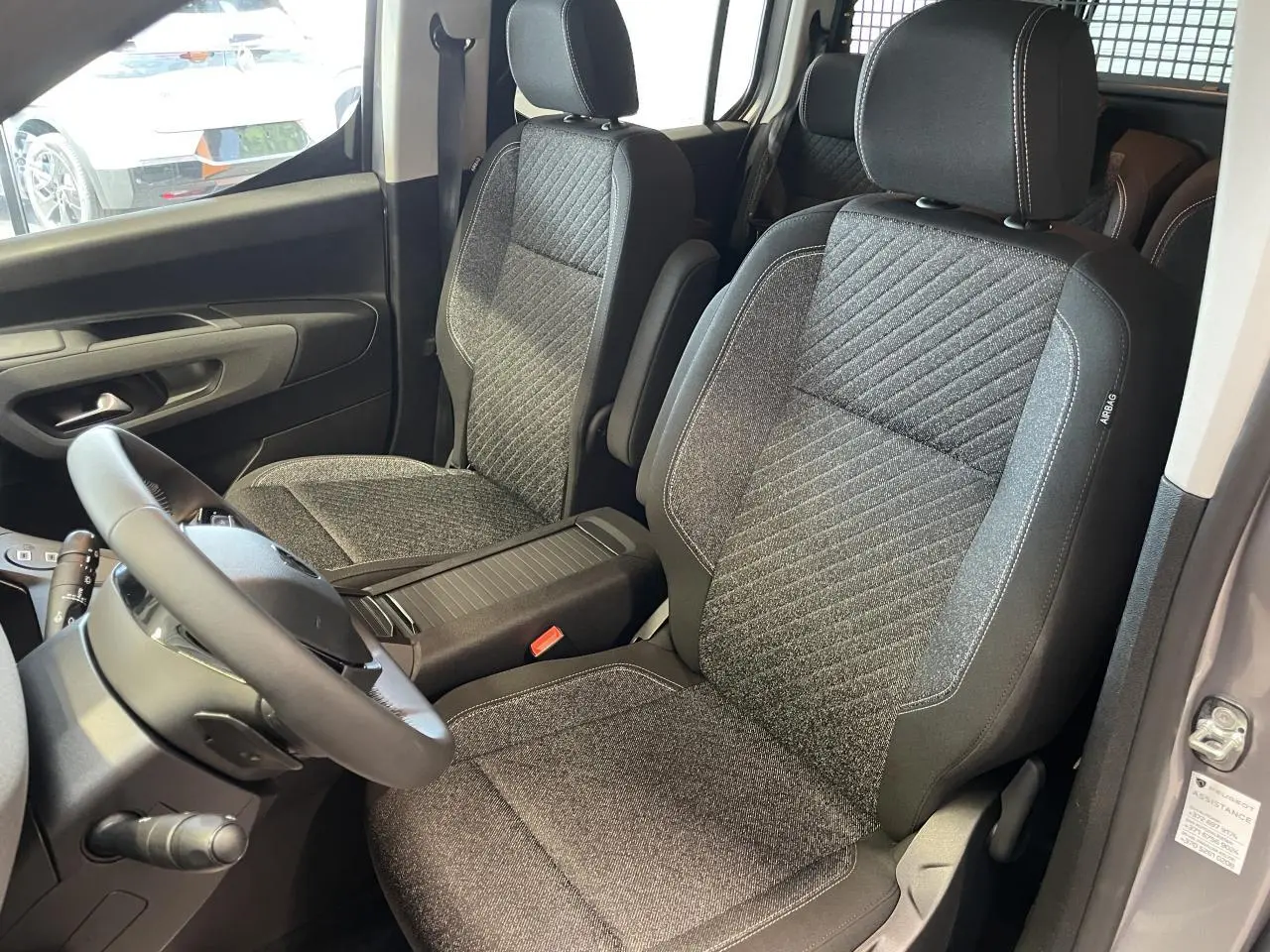 Vue intérieure sur les sièges avant en tissu gris du Peugeot Rifter 2026, avec volant cuir multifonction visible côté gauche.