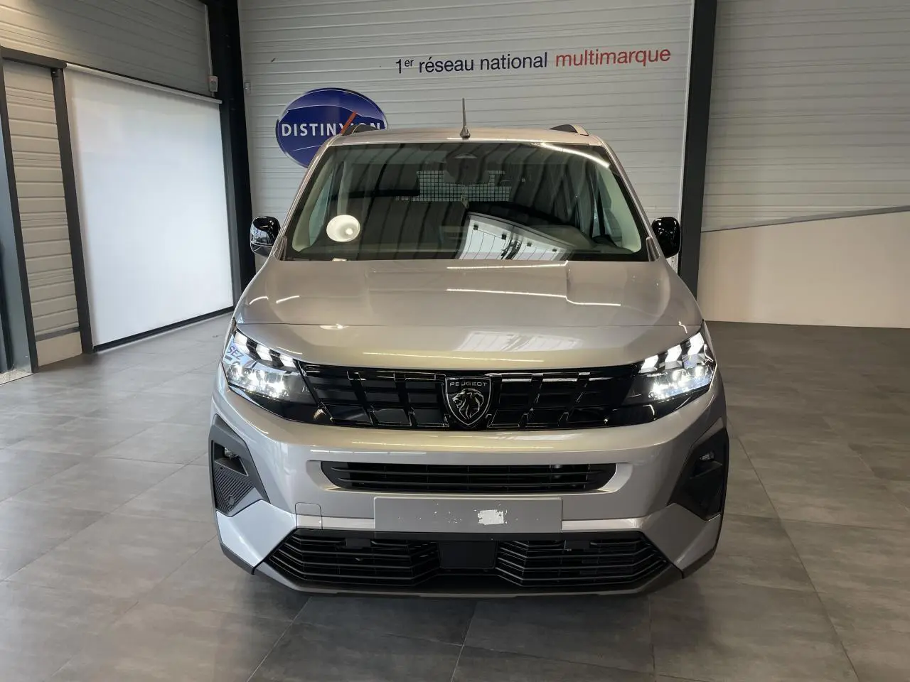 Vue de face du Peugeot Rifter gris Artense avec calandre noire et signature lumineuse LED allumée en intérieur showroom.