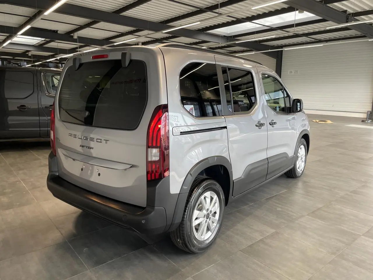 Peugeot Rifter gris Artense vu en 3/4 arrière droit dans un showroom, avec vitres teintées et feux verticaux LED.