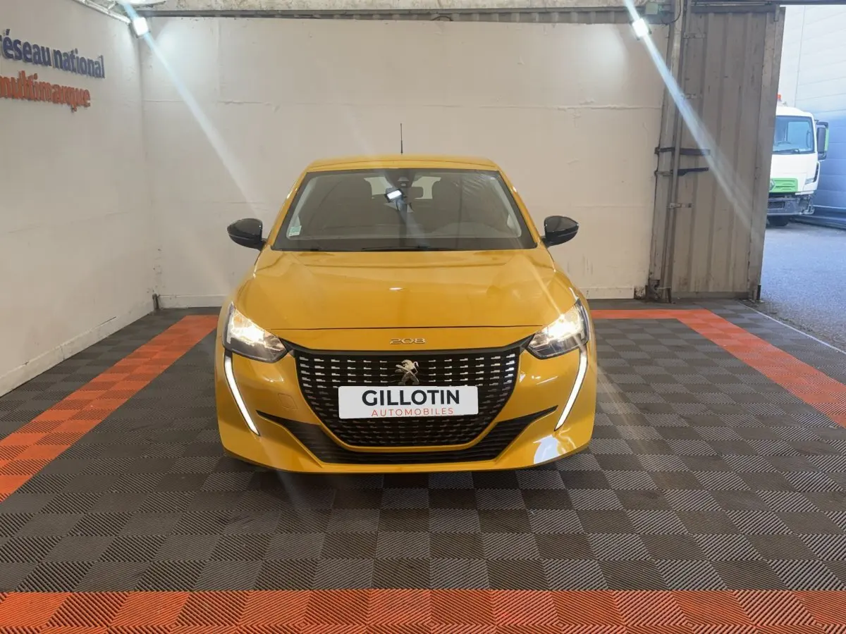 Vue frontale d'une Peugeot 208 jaune vif avec feux LED allumés, dans un espace intérieur de présentation.
