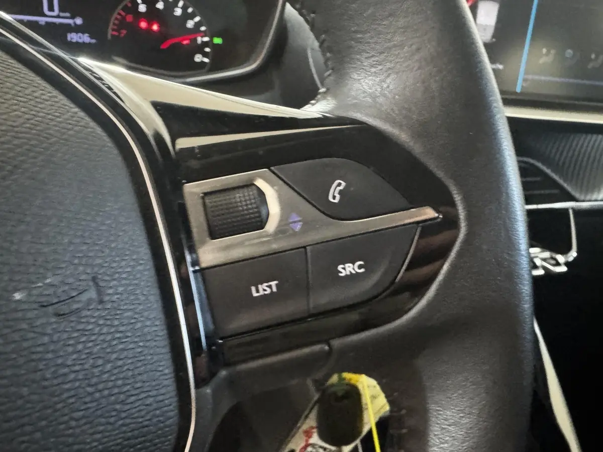Gros plan sur les commandes gauche du volant cuir noir d’une Peugeot 208 2023, avec tableau de bord en arrière-plan.