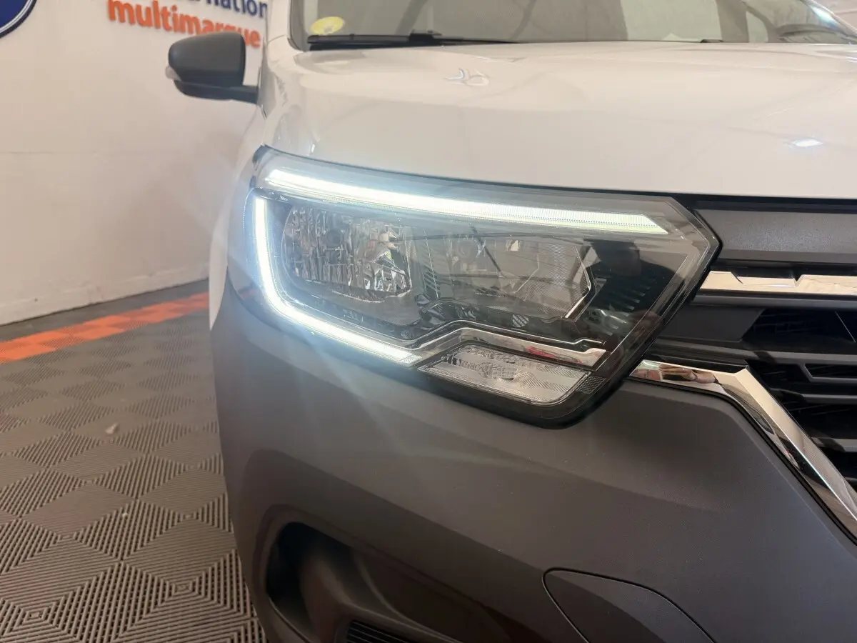 Gros plan sur le phare avant gauche allumé d’un Renault Kangoo blanc, soulignant la calandre noire et chromée.