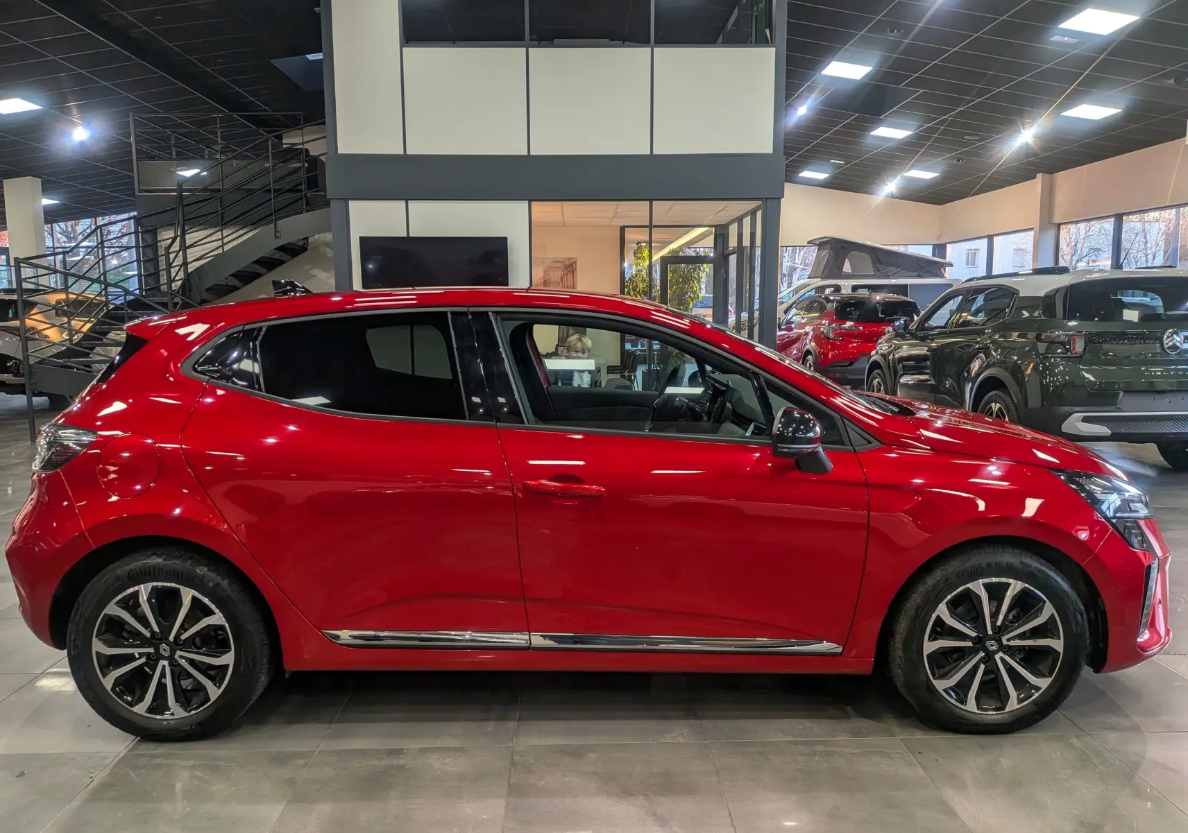 Profil latéral droit d'une Renault Clio V rouge flamme en showroom, avec jantes alliage et détails chromés sur les bas de porte.
