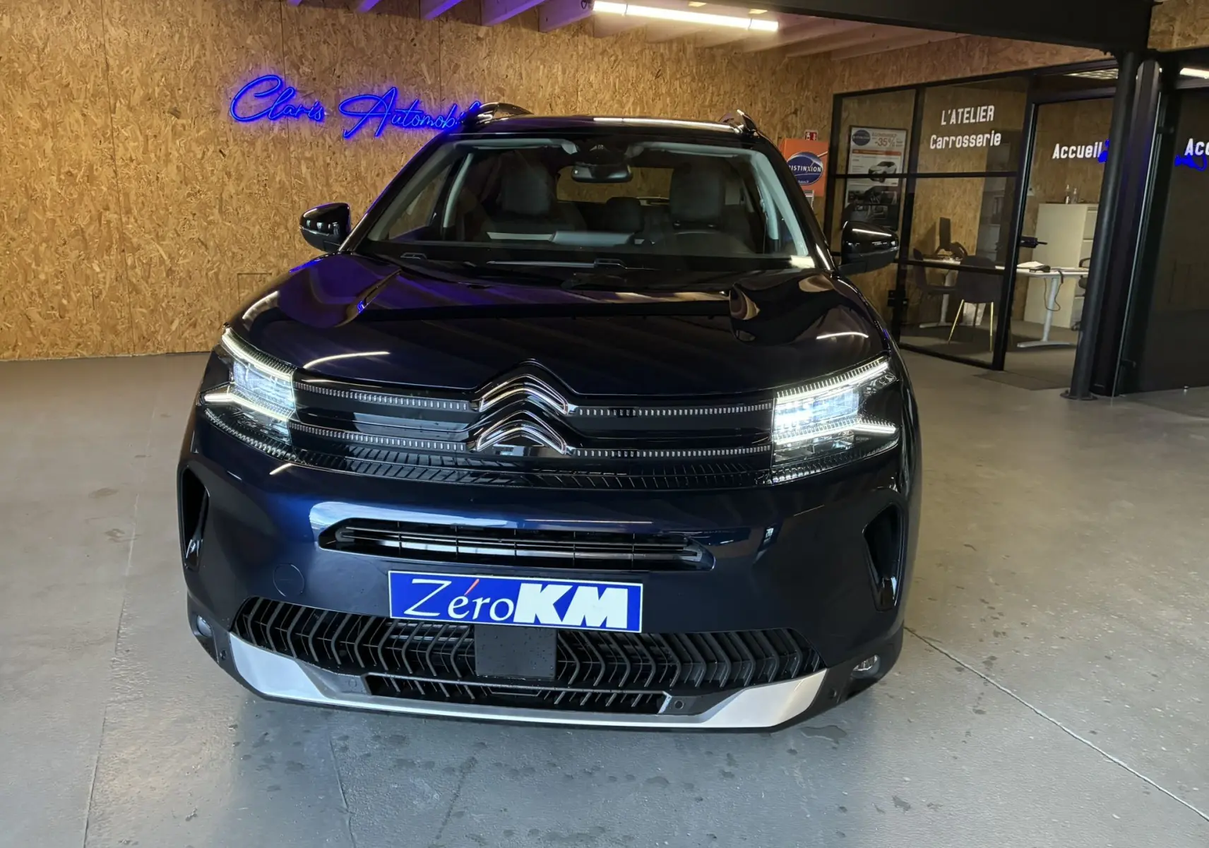 Vue avant d'un Citroën C5 Aircross bleu Eclipse Métal avec phares allumés et plaque "Zéro KM" en intérieur showroom.