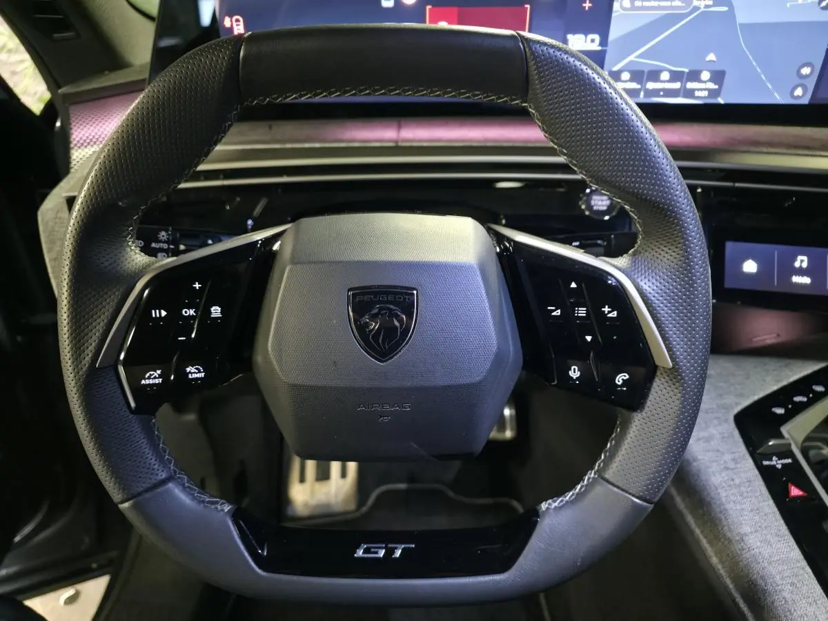 Gros plan sur le volant cuir perforé noir avec badge GT du Peugeot 3008 1.2i Hybrid 2025, tableau de bord numérique en arrière-plan.