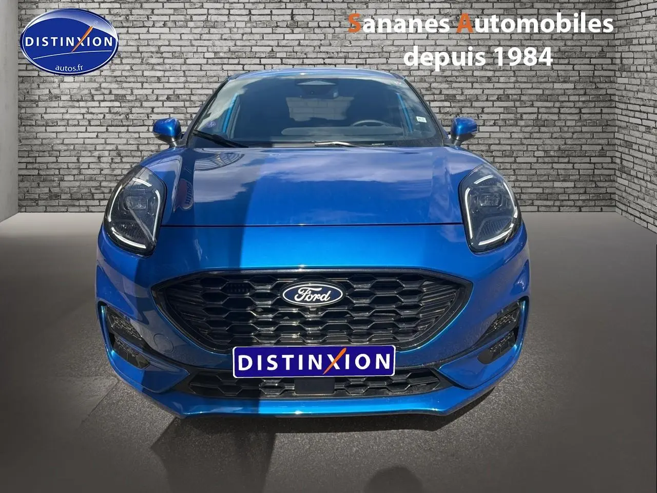 Vue frontale d'une Ford Puma bleu métallisé avec calandre noire et phares LED, sur fond de mur en briques grises.
