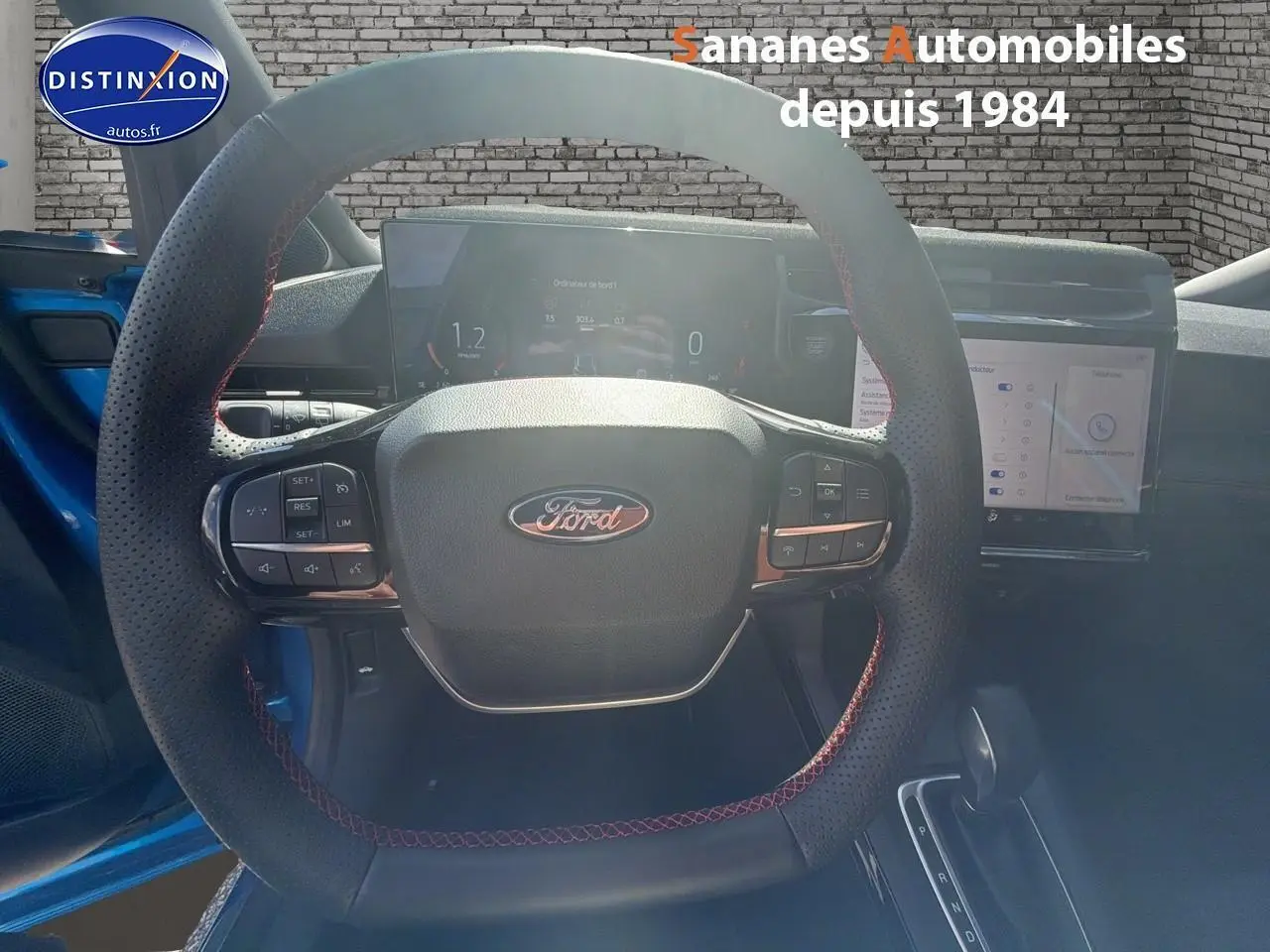 Vue intérieure centrée sur le volant perforé avec surpiqûres rouges du Ford Puma bleu 2025, tableau de bord numérique et écran tactile visibles.