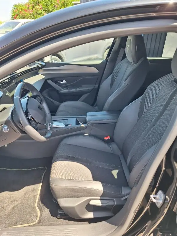 Intérieur avant droit de la Peugeot 308 Hybrid 145 GT 2025 noire, sièges tissu et cuir avec accoudoir central.