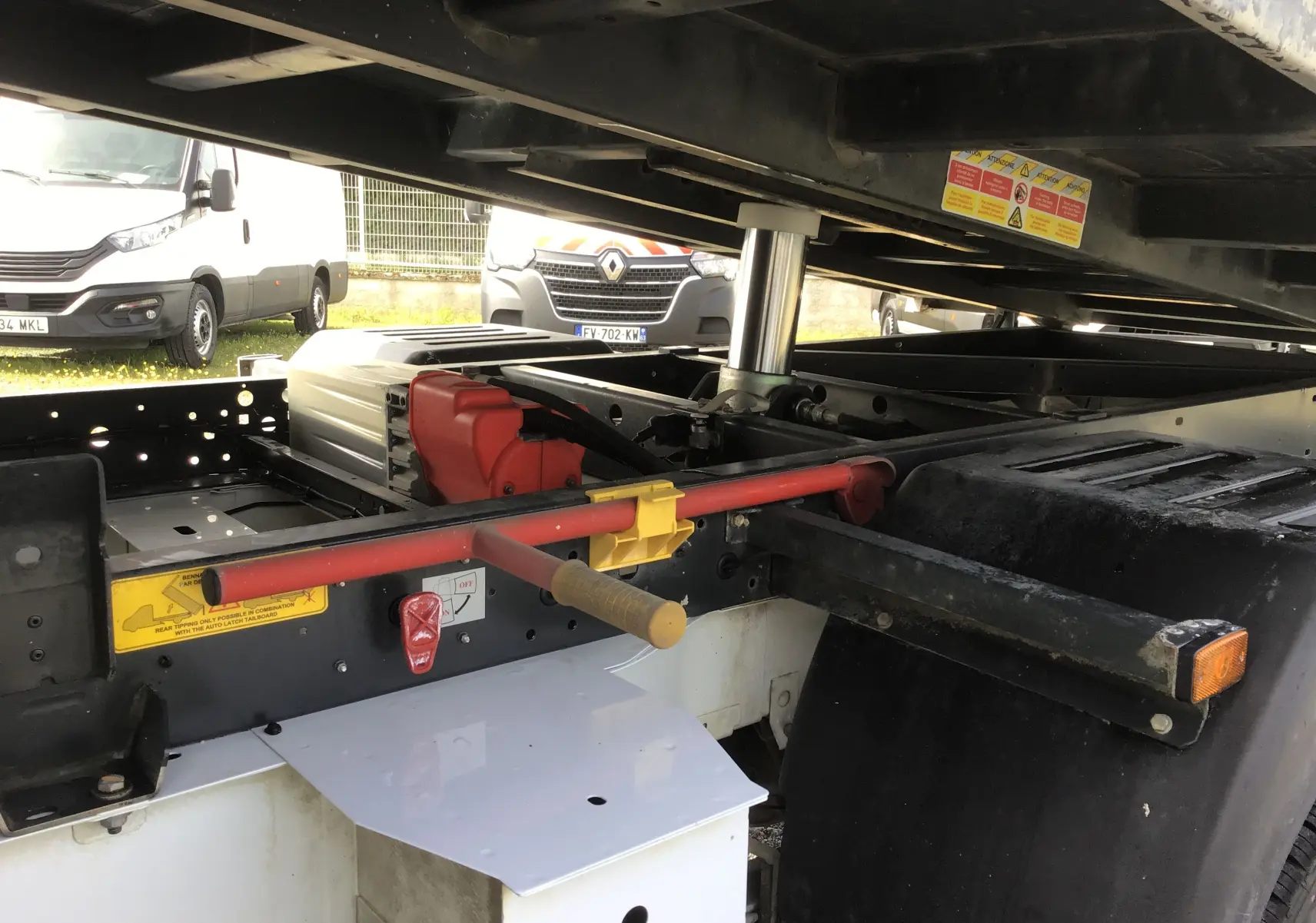 Vue rapprochée sous la benne relevée d’un Renault Master blanc, montrant le châssis, vérin hydraulique et coffre latéral.