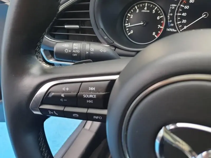 Gros plan sur le volant cuir noir de la Mazda3 2025 avec commandes audio et compteur partiel en arrière-plan.