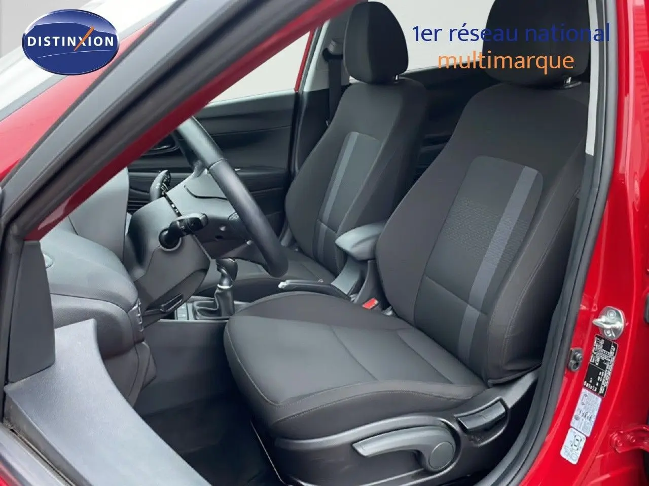 Vue intérieure côté conducteur de la Hyundai i20 rouge Dragon Metal 2025, sièges tissu noir et volant cuir multifonction.