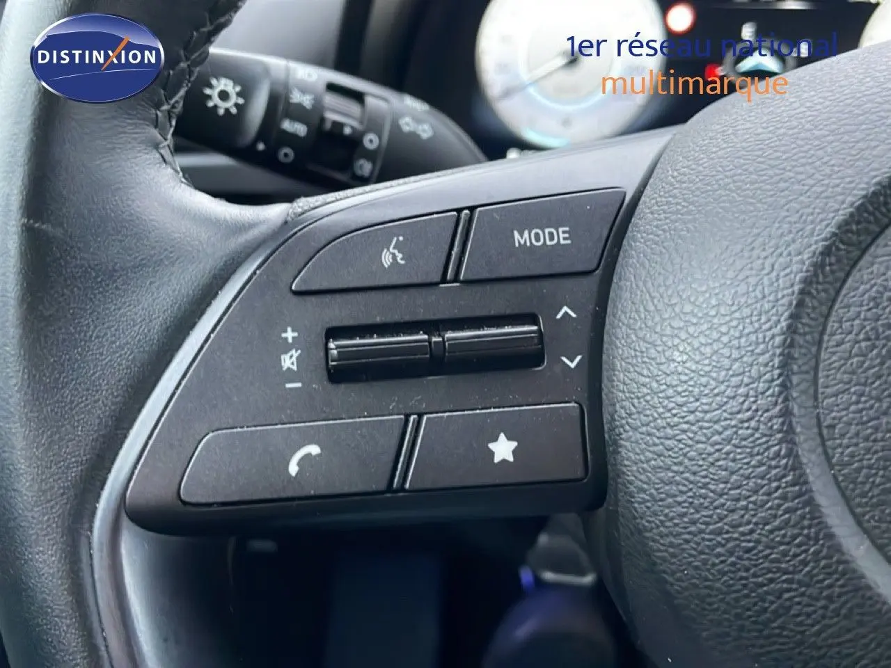 Gros plan sur les commandes gauche du volant en cuir noir de la Hyundai i20 Rouge Dragon Metal 2025, avec tableau de bord flou en arrière-plan.