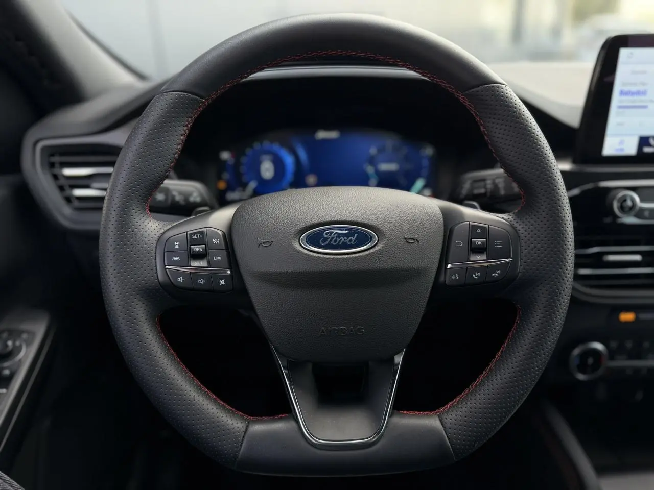 Gros plan sur le volant noir perforé avec surpiqûres rouges du Ford Kuga 2023, tableau de bord numérique en arrière-plan.