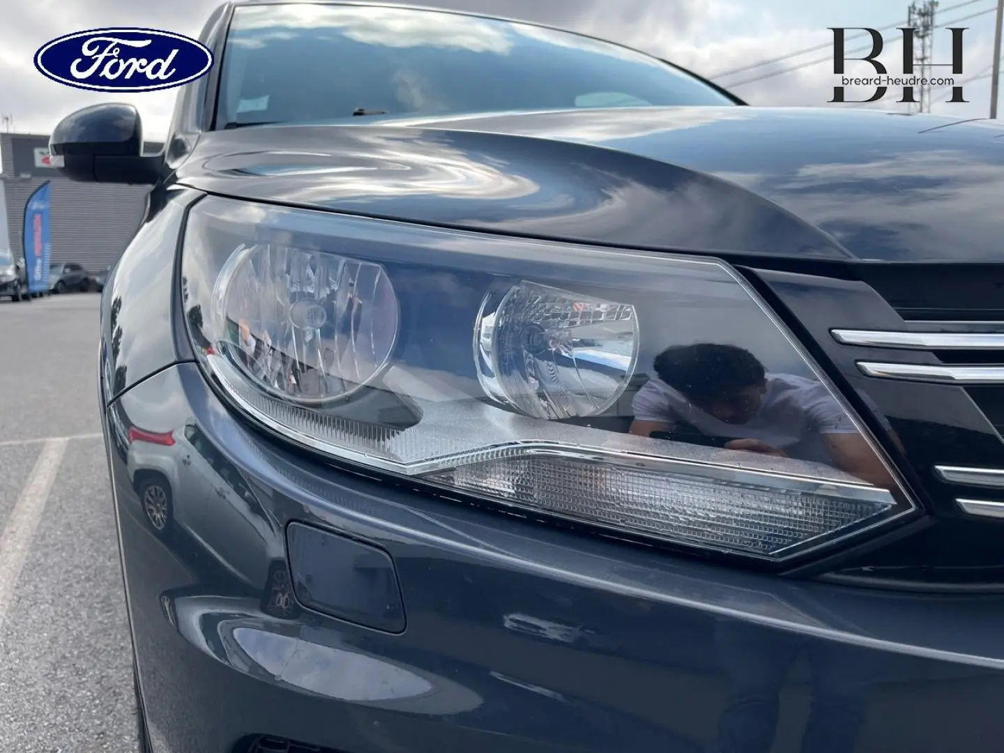 Gros plan sur le phare avant gauche d’un Volkswagen Tiguan gris poivre avec calandre chromée visible.