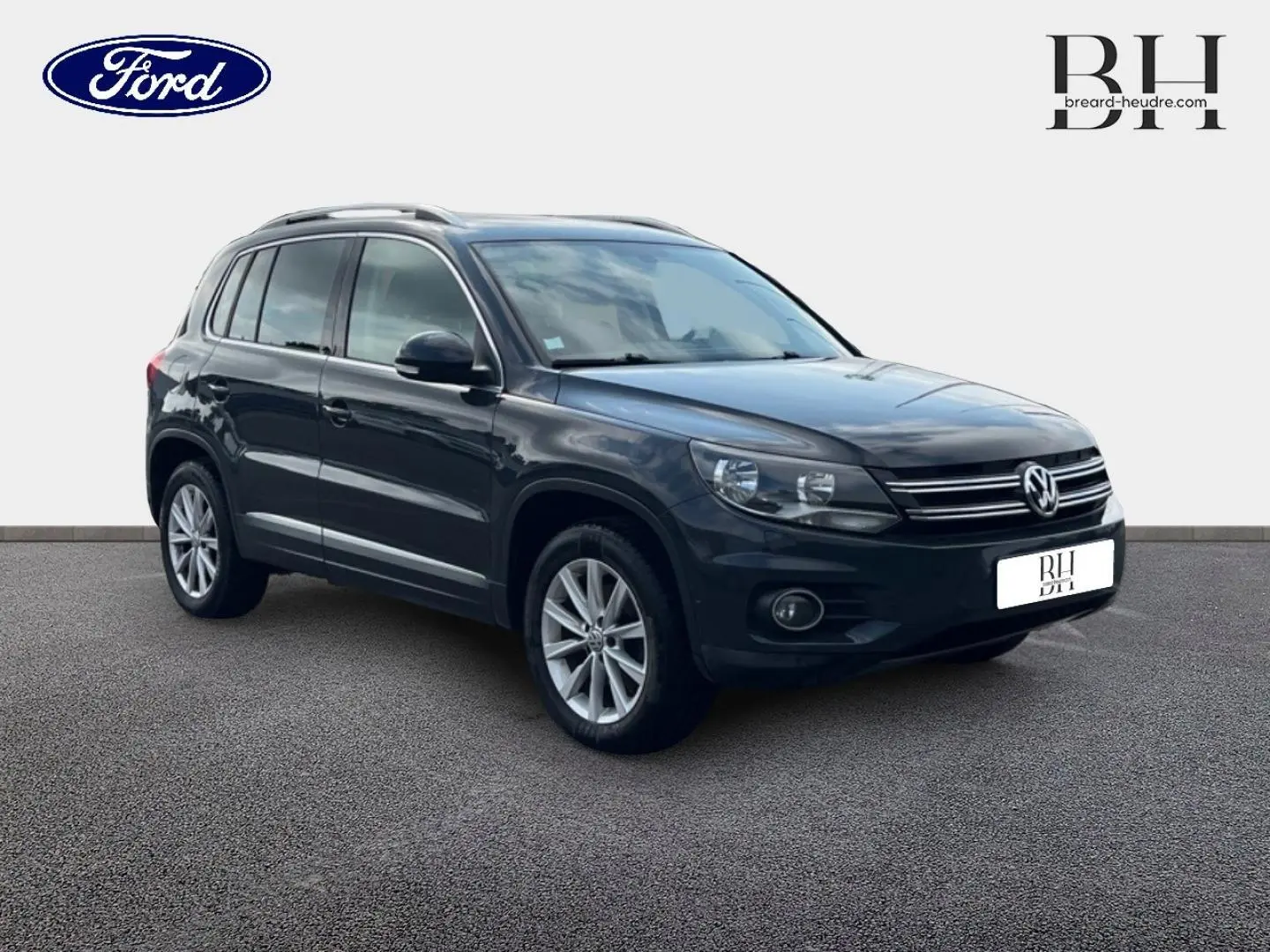 Volkswagen Tiguan gris poivre en 3/4 avant droit, avec jantes alu et barres de toit visibles.