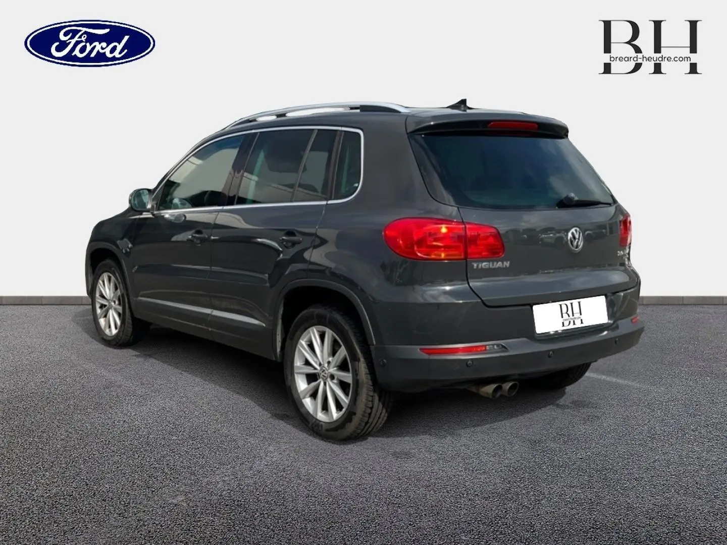 Vue 3/4 arrière droite d'un Volkswagen Tiguan gris poivre avec jantes alu et barres de toit visibles.