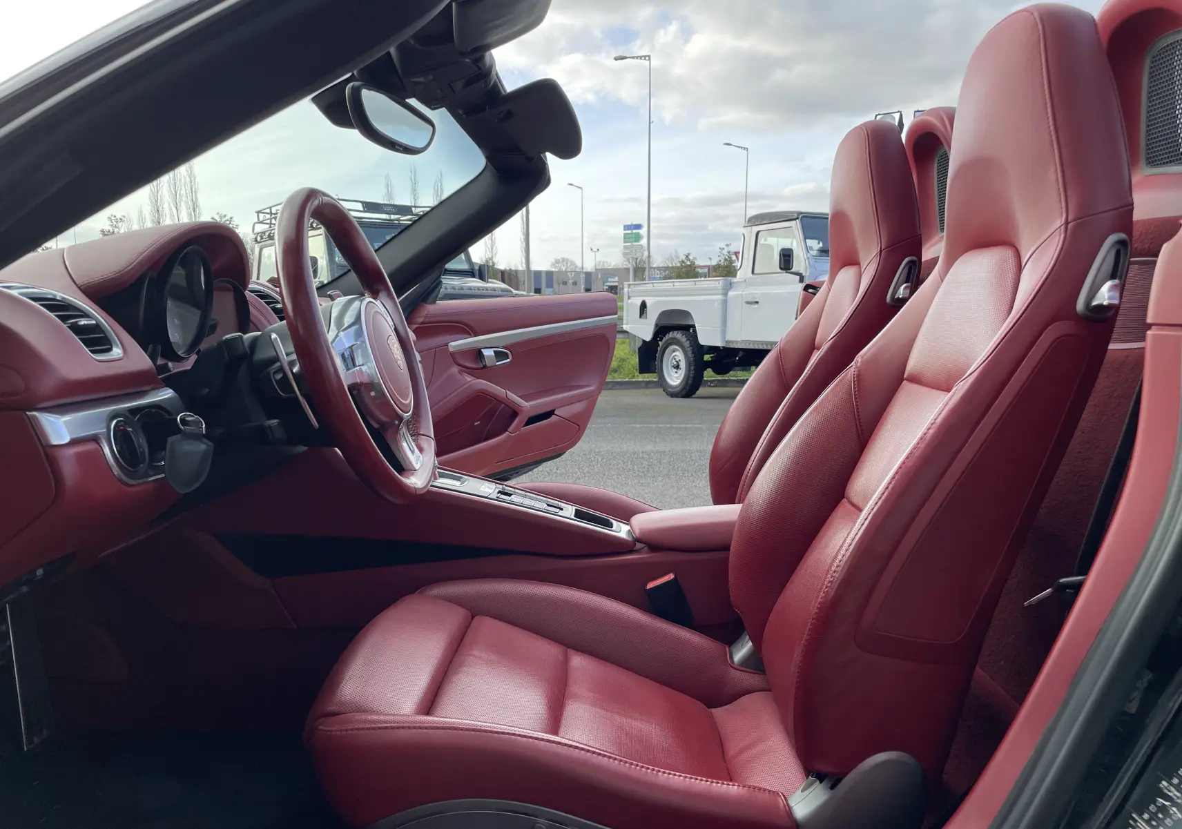 Intérieur cuir rouge grenat vu côté gauche d'une Porsche Boxster S 981 noire, volant SportDesign et console centrale visibles.