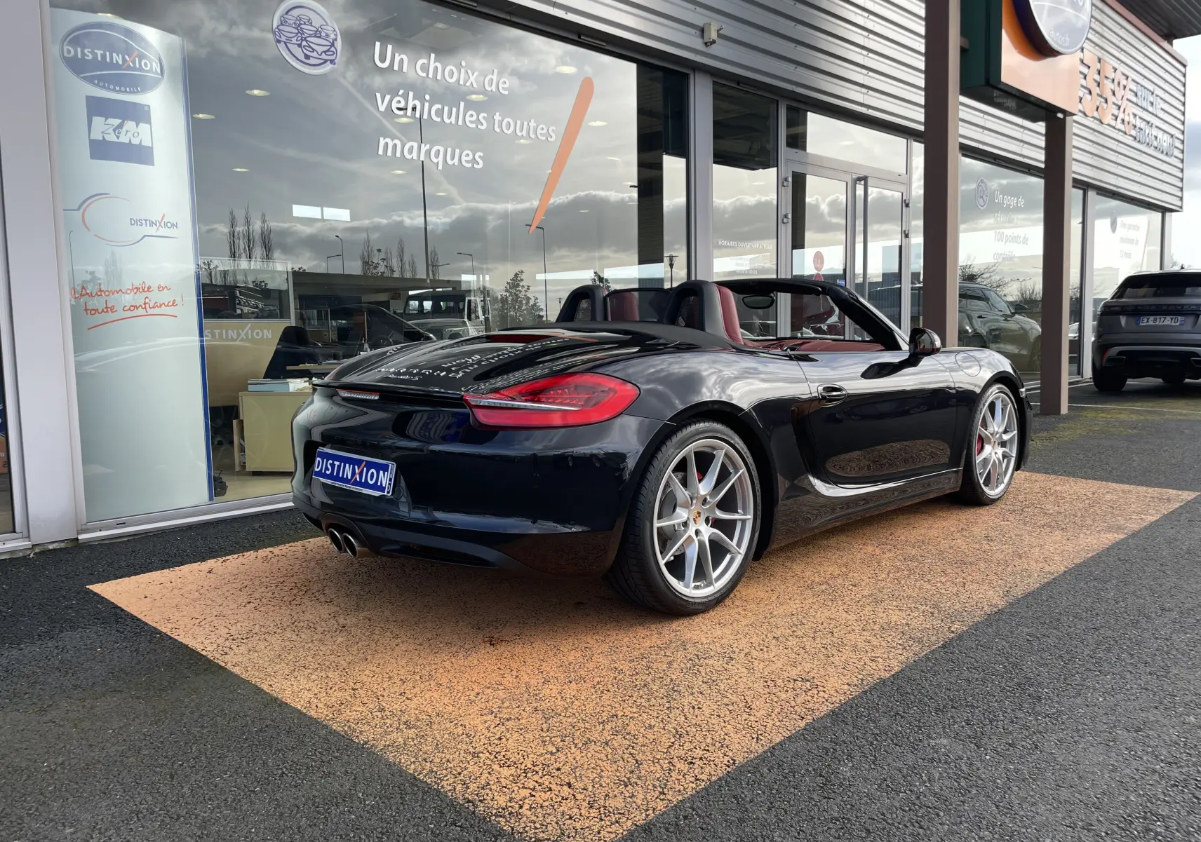 Vue 3/4 arrière droite d'une Porsche Boxster S noire intense cabriolet, avec jantes argentées et intérieur cuir rouge grenat visible.