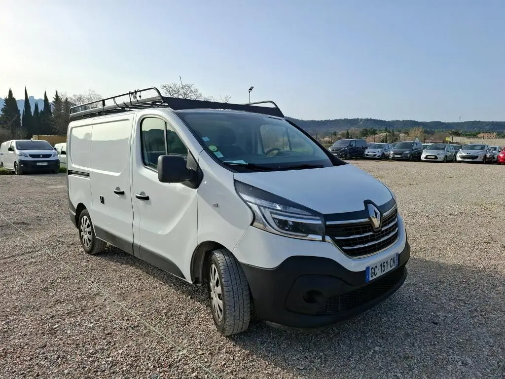 Fourgon Renault Trafic blanc vu en 3/4 avant droit avec galerie de toit, stationné sur un sol gravillonné.