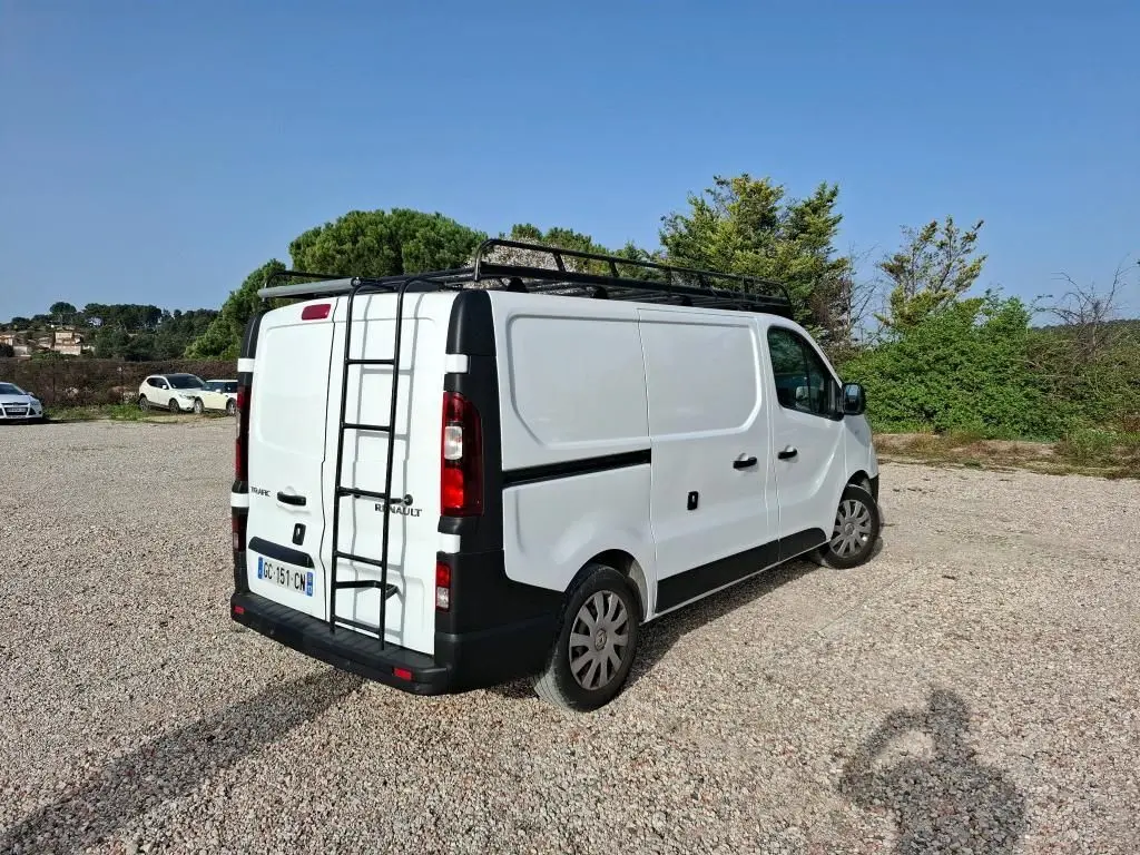 Renault Trafic Fourgon blanc vu en 3/4 arrière droit avec galerie et échelle sur le toit, stationné sur un sol gravillonné.