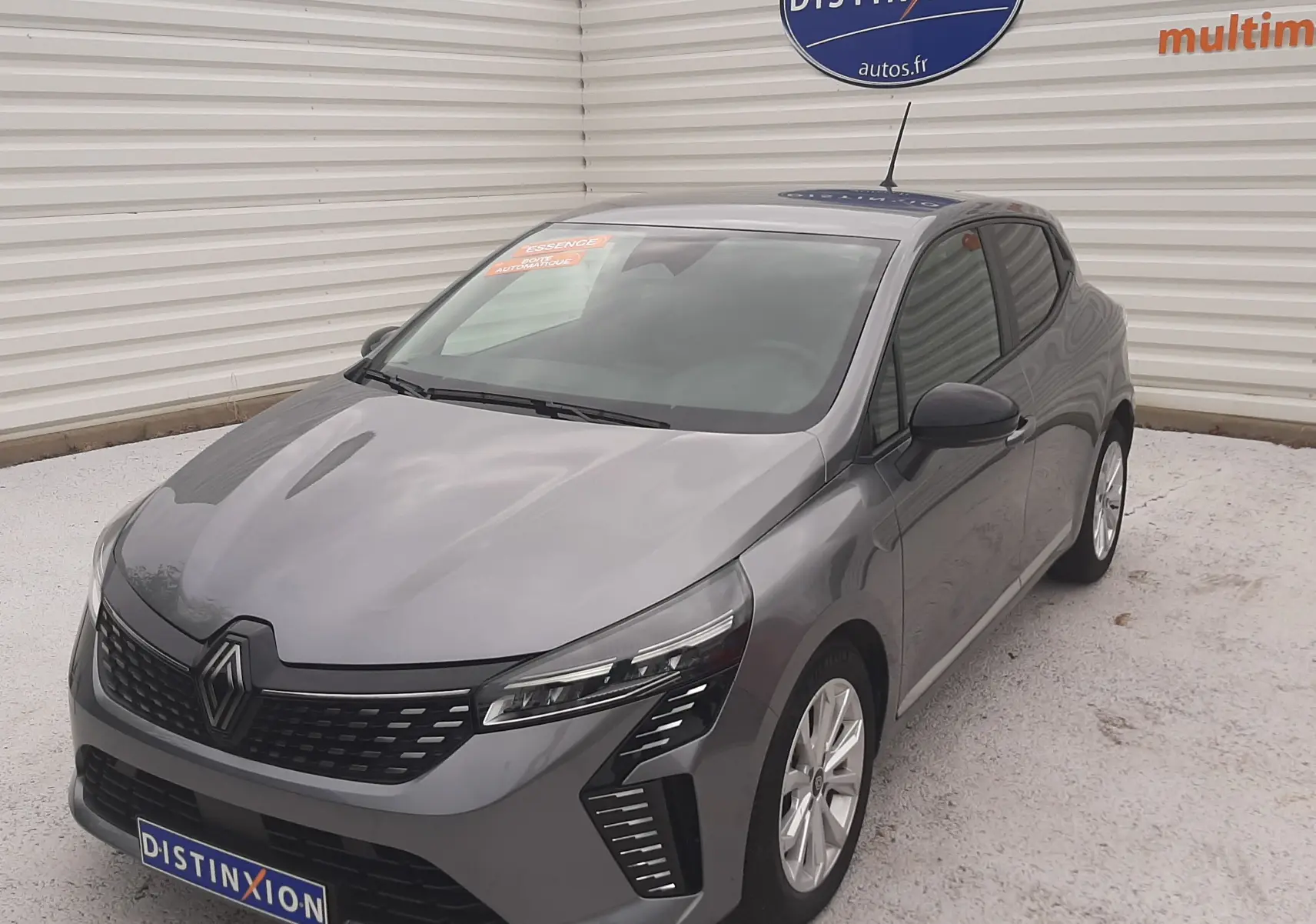 Renault Clio V gris schiste vue 3/4 avant droit, avec calandre noire et feux LED distinctifs, sur parking extérieur.