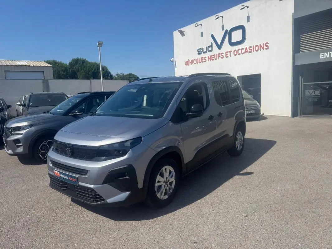 Peugeot Rifter gris en 3/4 avant droit, avec barres de toit et jantes alliage, stationné devant un bâtiment blanc.