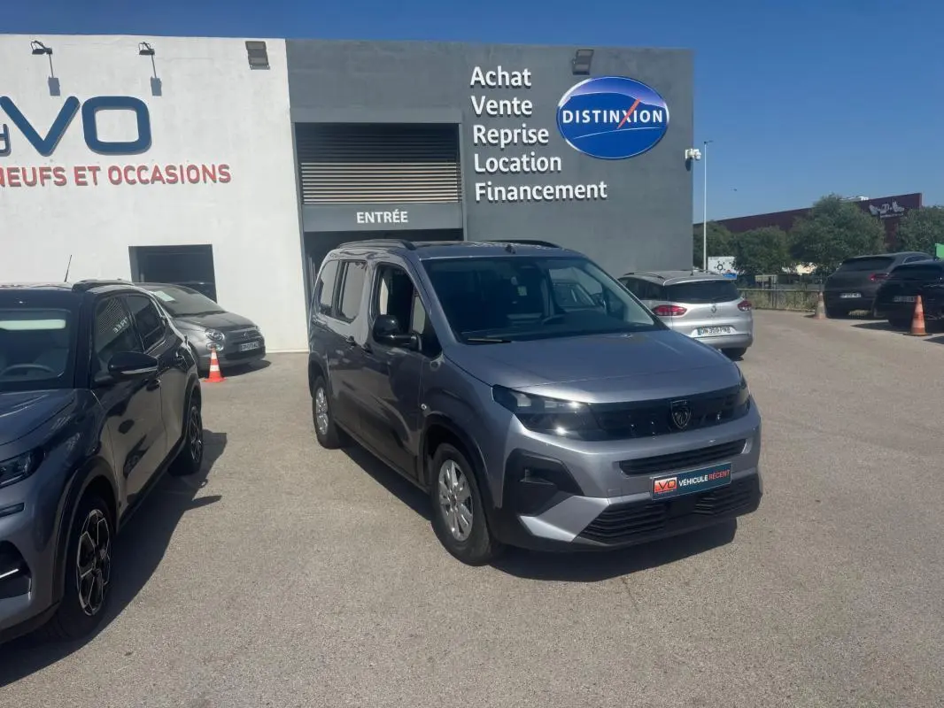 Peugeot Rifter gris vue 3/4 avant droit, avec barres de toit et jantes alliage, stationné devant un bâtiment commercial.