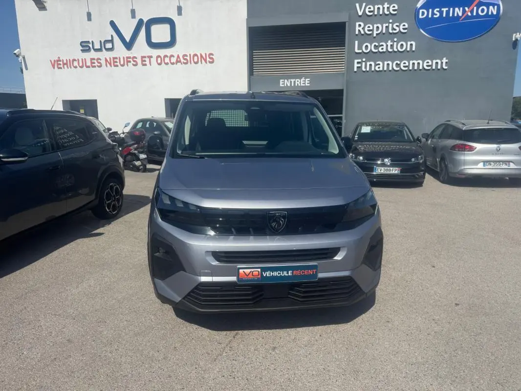 Vue avant d’un Peugeot Rifter gris 2026 avec calandre noire et logo Peugeot visible, stationné devant un concessionnaire.