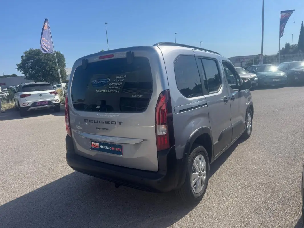 Peugeot Rifter gris vue 3/4 arrière droit, avec barres de toit et vitres teintées, stationné en extérieur.
