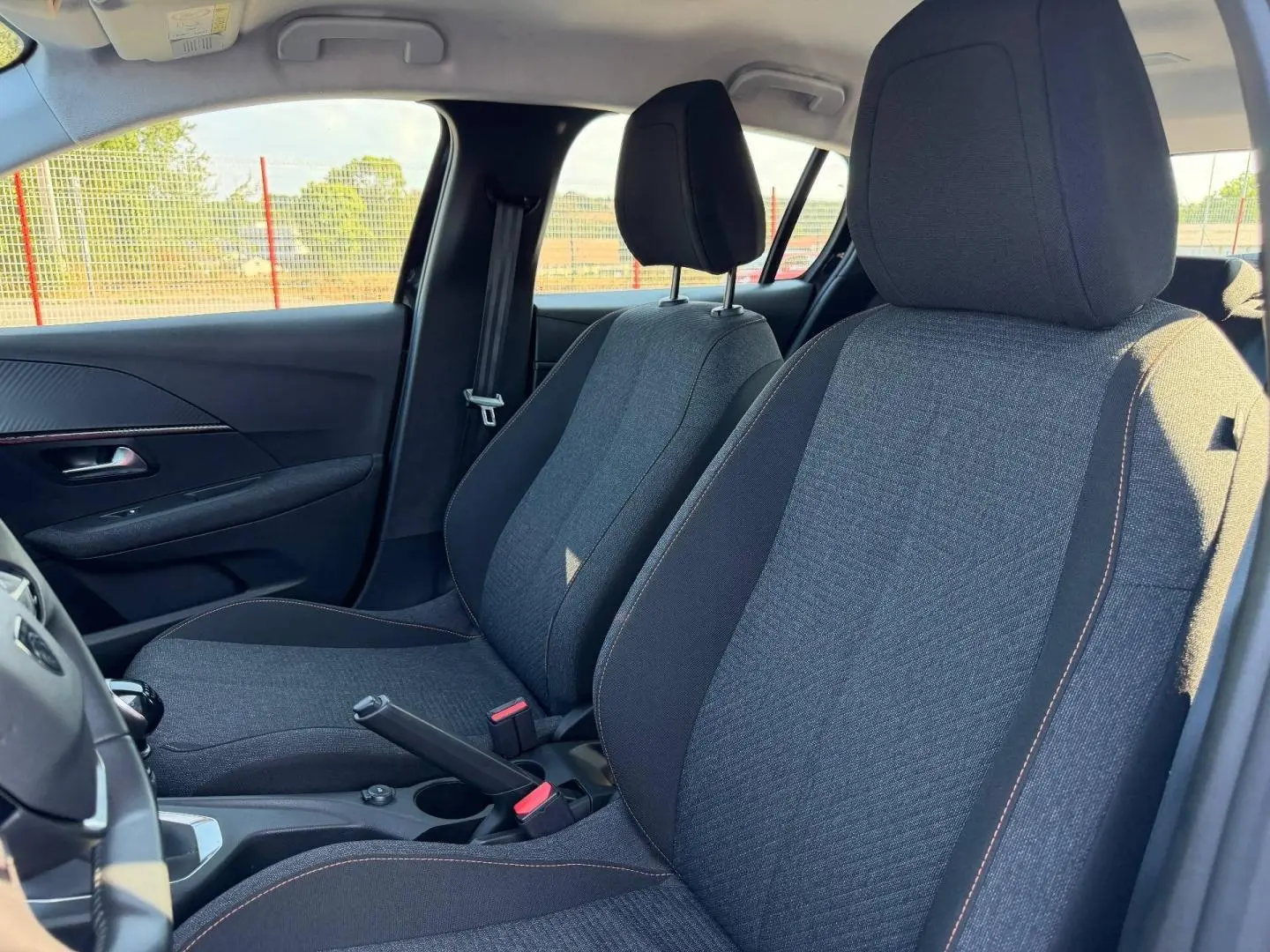 Vue intérieure côté gauche montrant les sièges avant en tissu gris avec surpiqûres orange de la Peugeot 208 2025.