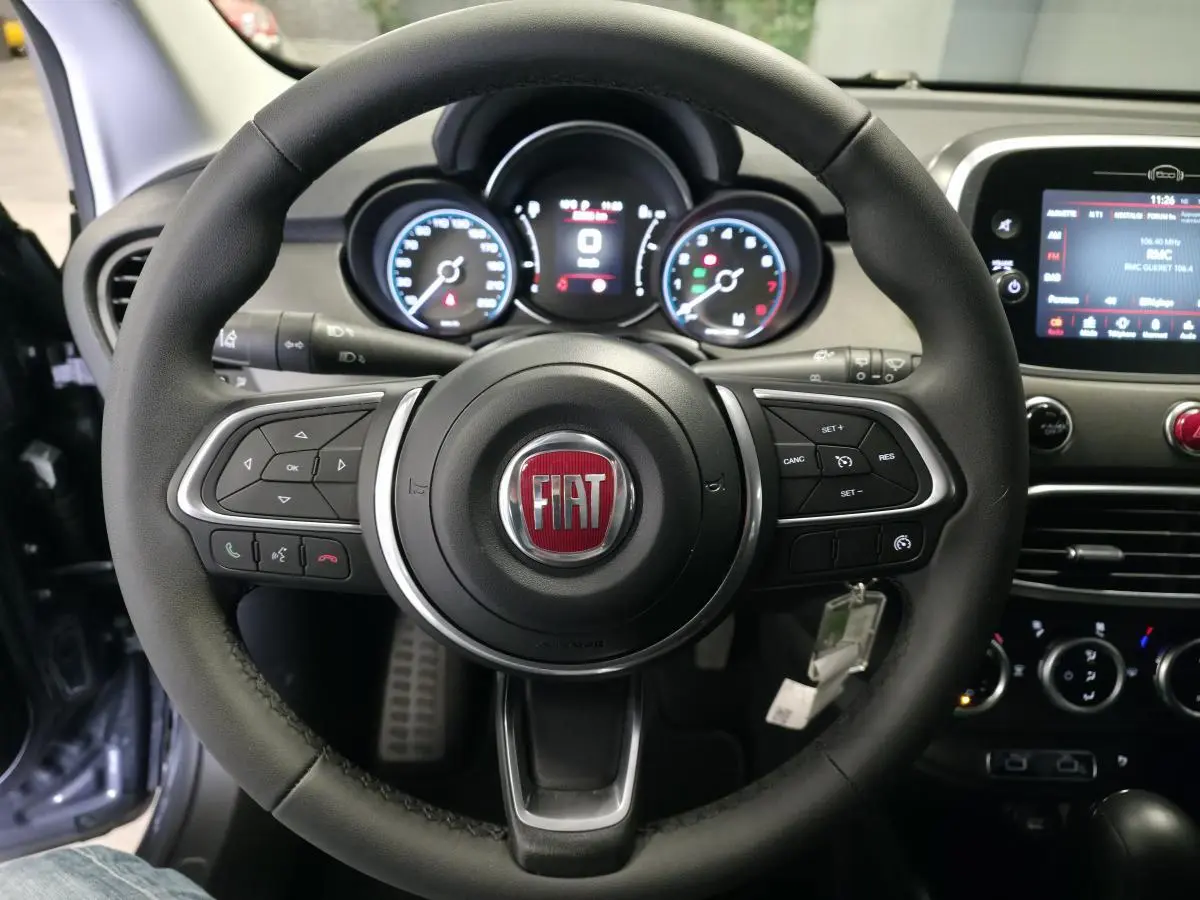 Volant en cuir noir avec logo FIAT au centre, tableau de bord digital et écran tactile du Fiat 500X gris, vue de face intérieure.