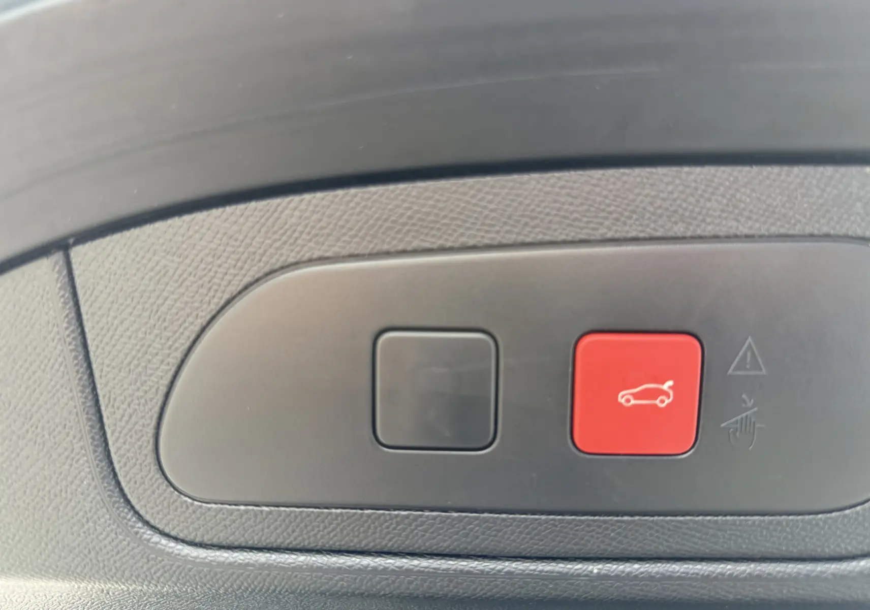Boutons de commande du coffre, dont un rouge avec pictogramme de voiture, sur panneau intérieur noir texturé.