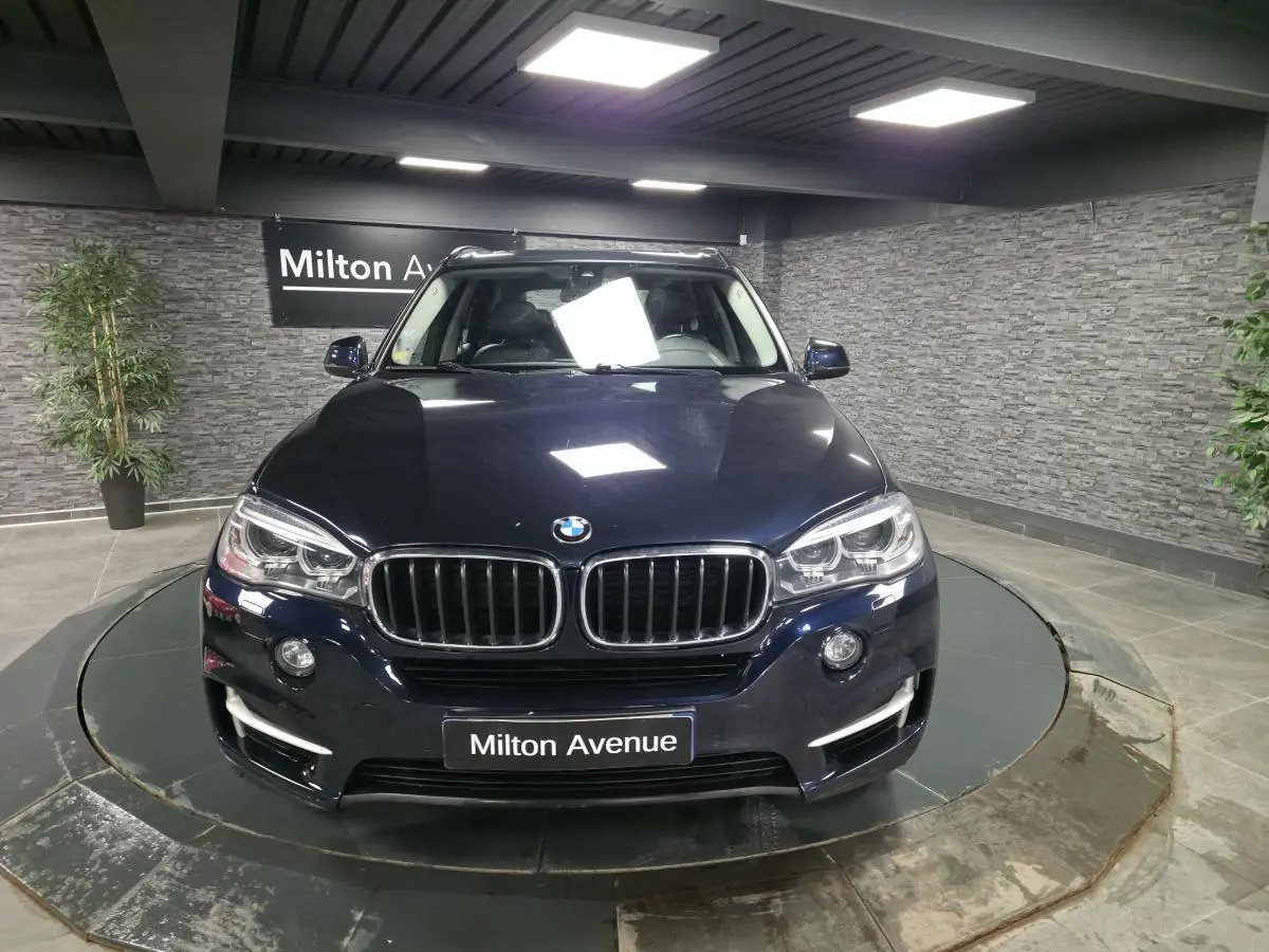 BMW X5 sDrive 25d bleu métallisé vue de face avec calandre noire et projecteurs bi-xénon allumés.