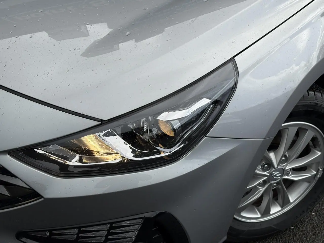 Gros plan sur le phare avant droit et la jante alliage d'une Hyundai i30 SW T-GDI 120 Modern gris argent sous la pluie.