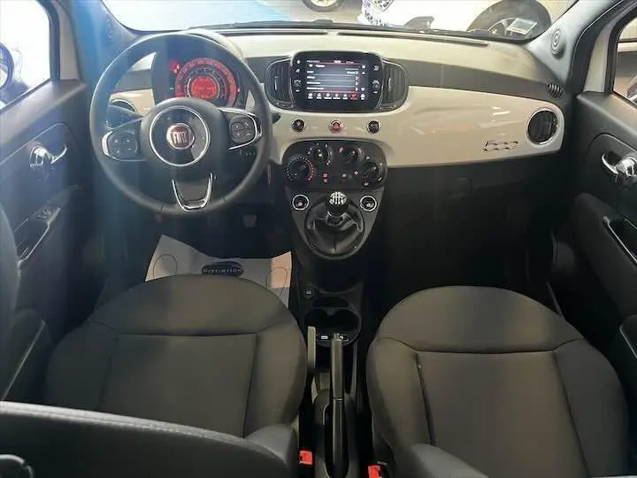 Intérieur avant de la Fiat 500 blanche 2023, tableau de bord clair avec écran tactile et volant multifonction noir.