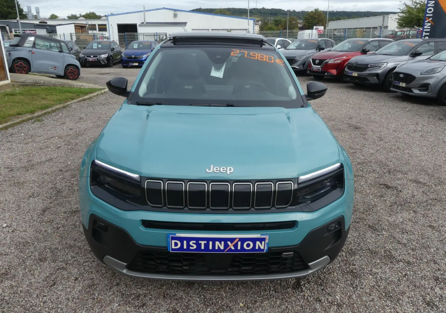 Vue frontale d'un Jeep Avenger bleu métallisé avec calandre noire et plaque Distinxion sur un parking.